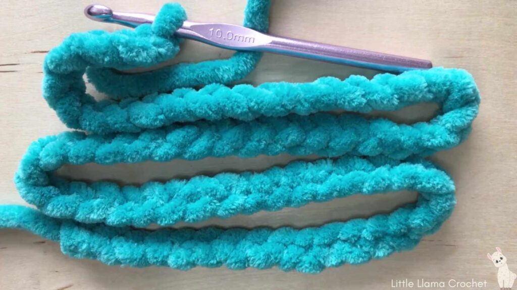 A flat lay on a light wooden background of a thick foundation chain in blue chenille yarn laying back and forth on itself with a 10 mm aluminum crochet hook at the top.