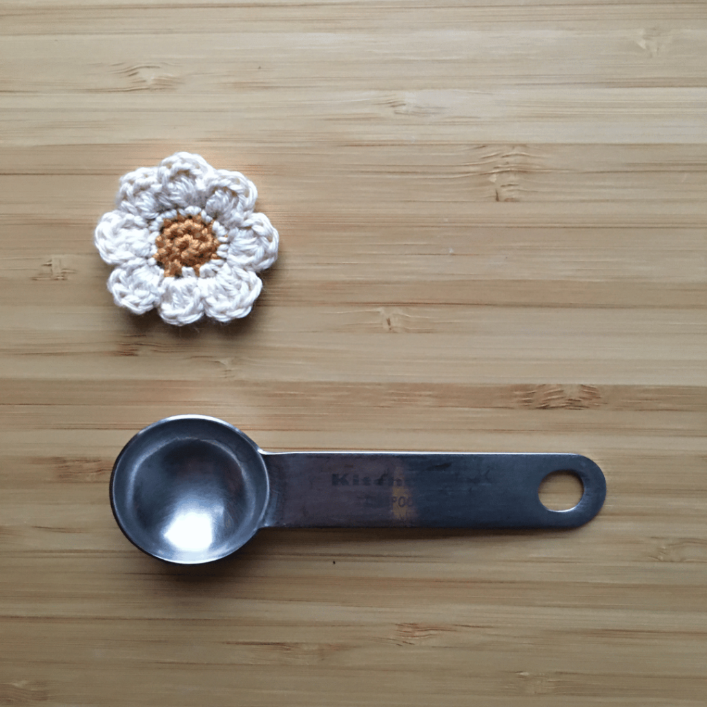 A small white and gold crochet flower lays beside a metal teaspoon both are on a wooden background. 