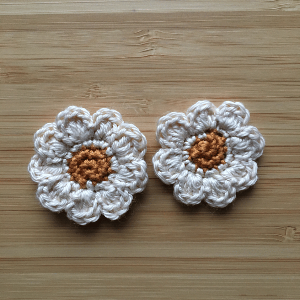 Flat lay of two small dainty crochet daisies on a light wooden background. The centre of the crochet daisy is golden cotton and the petals are made out of cream coloured cotton.