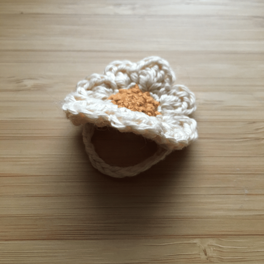 top profile of a small dainty crochet daisy flower ring on a light wooden background. The centre of the crochet daisy is gold and the petals are made out of cream coloured cotton.