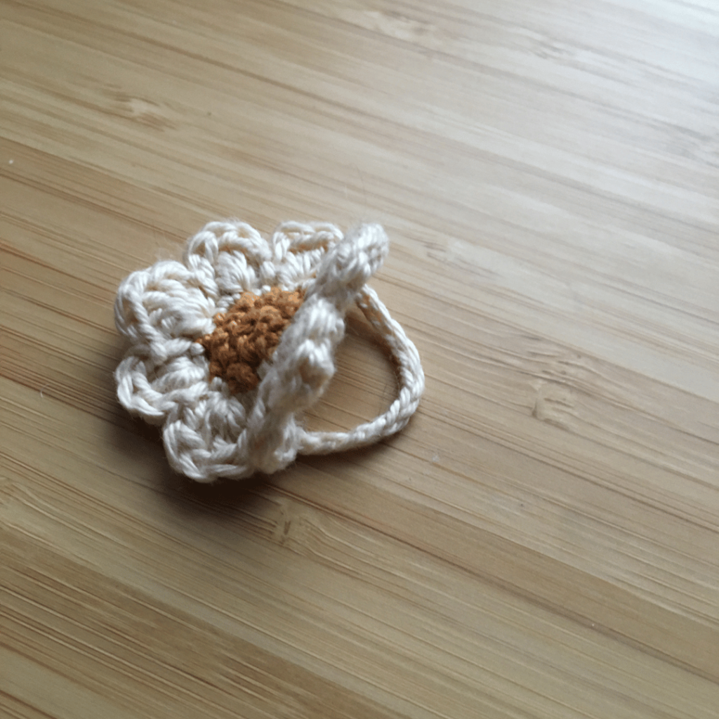 Side profile of a small dainty crochet daisy flower ring on a light wooden background. The centre of the crochet daisy is gold and the petals are made out of cream coloured cotton.