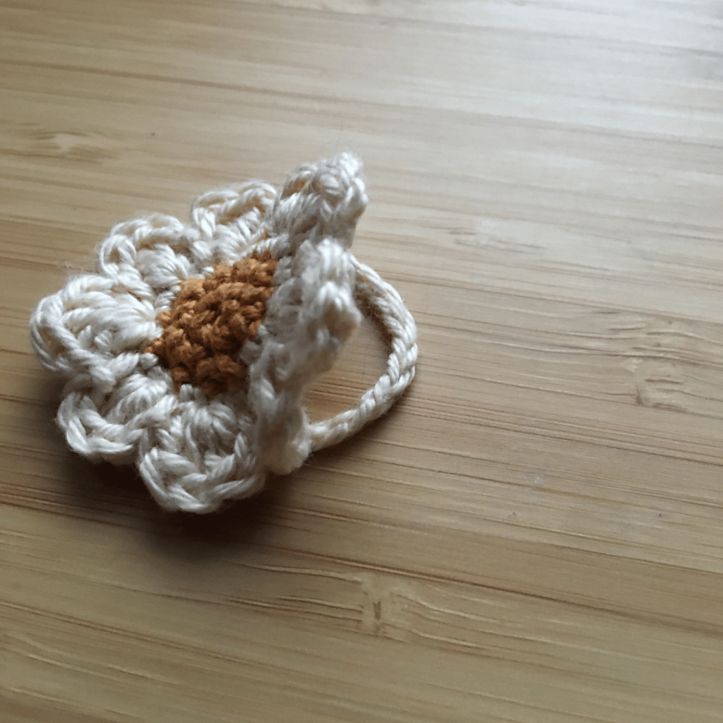 Side profile of a small dainty crochet daisy flower ring on a light wooden background. The centre of the crochet daisy is gold and the petals are made out of cream coloured cotton.