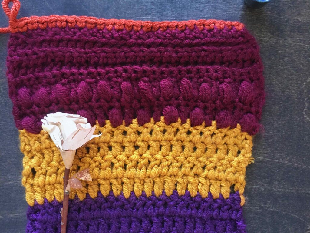 Vertical crochet flat lay on dark wood featuring a stitch swatch of  maroon crochet puff stitches at the top and gold and purple double crochet below. A birch rose lays on the left side of the crochet fabric. 