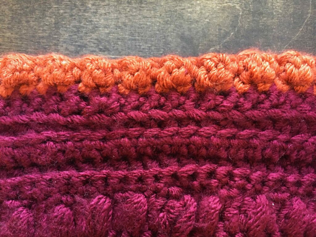 Vertical crochet flat lay on a background of dark wood of a stitch sampler with a row of orange bobble stitches across the top and maroon puff stitches below. 