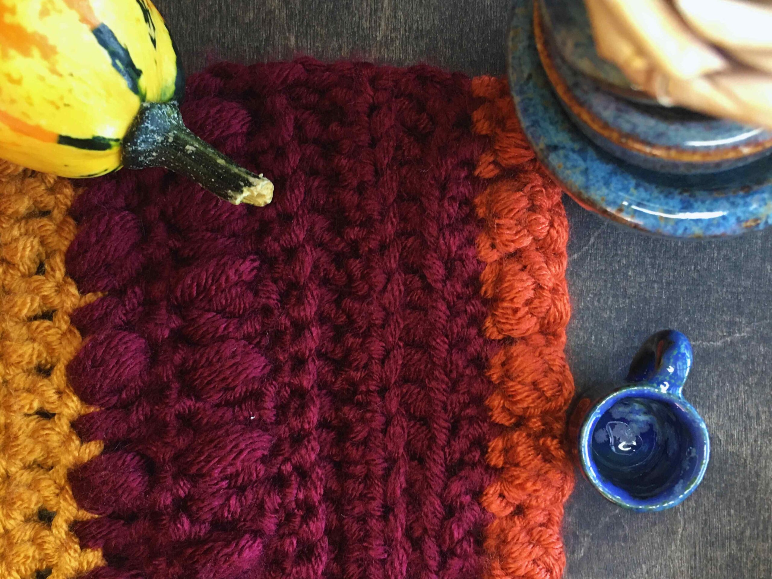 Crochet flat lay with a row of maroon puff stitches and orange bobble stitches. A tiny blue cup sits in the bottom right corner. A birch rose sits in the top right corner of the picture and an orange gourd with a green stem lays in the top left corner.