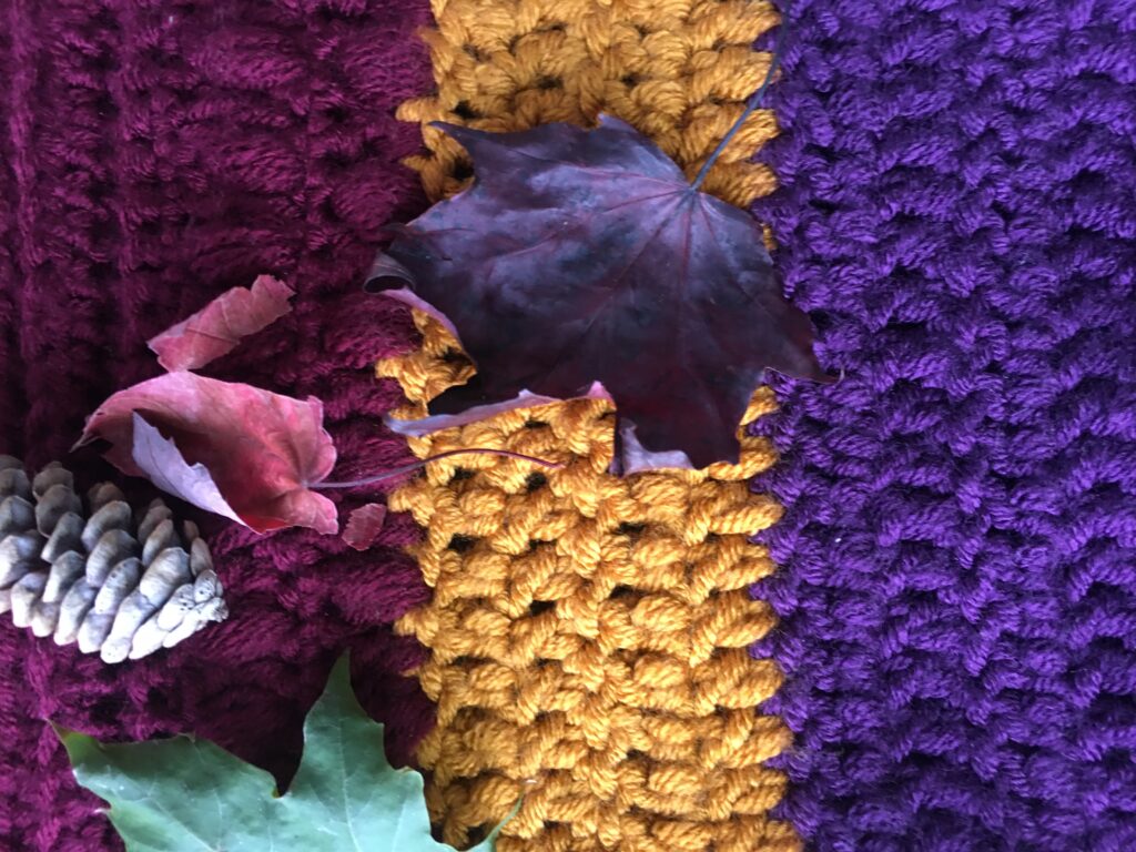 Crochet fabric featuring the double crochet lay flat in maroon, gold and purple. Fall leaves and a pinecone are on top on the fabric. 