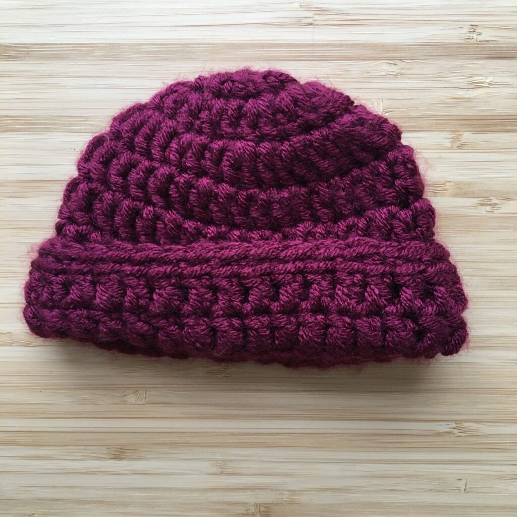 A maroon completed crochet hat for a newborn rests on a light wooden background