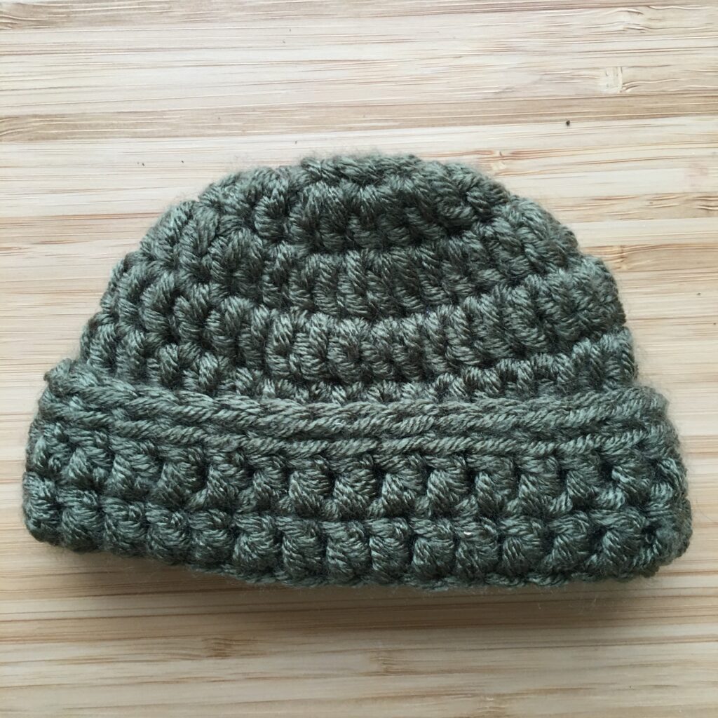 A green crochet newborn hat with a cap and brim rests completed on a light wooden background. 