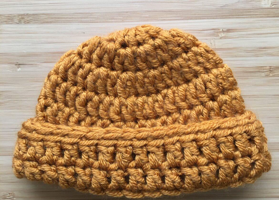 A gold crochet newborn hat rests on a light wooden background, the newborn cap has a folded brim made of double crochet