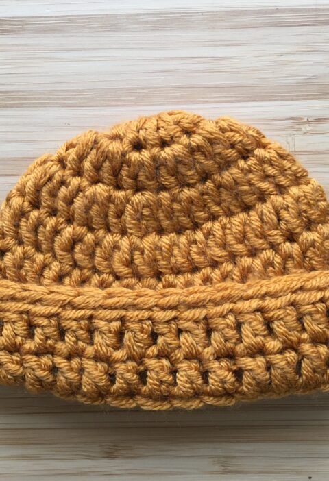 A gold crochet newborn hat rests on a light wooden background, the newborn cap has a folded brim made of double crochet