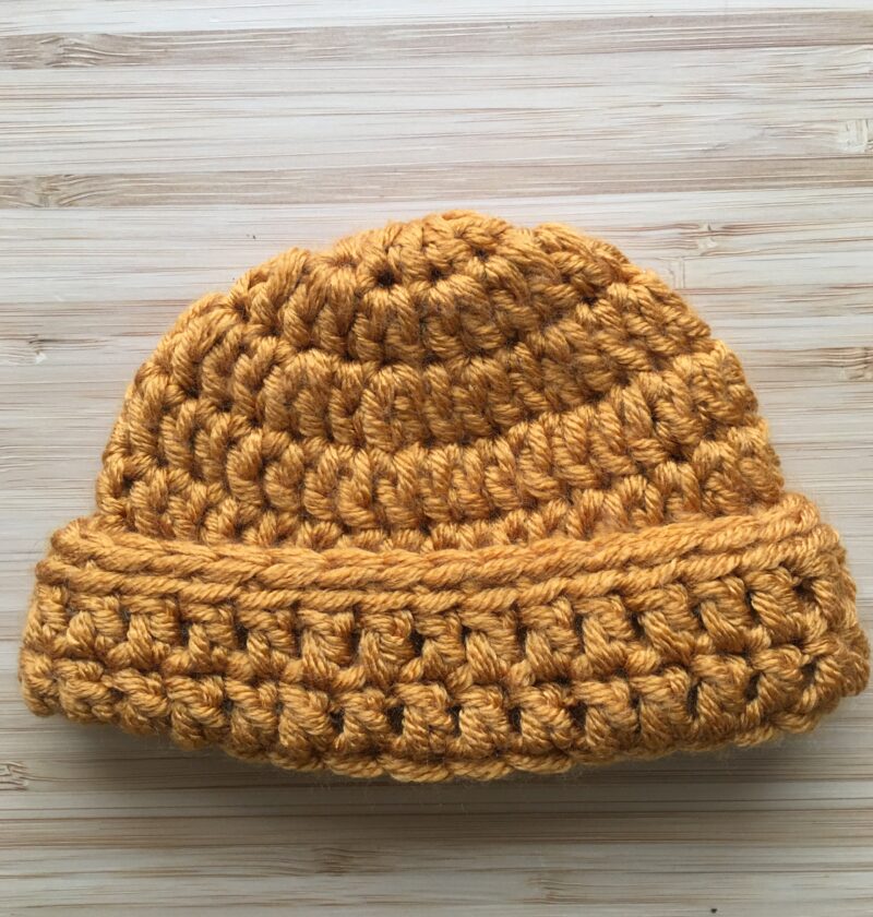 A gold crochet newborn hat rests on a light wooden background, the newborn cap has a folded brim made of double crochet