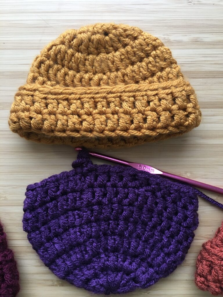 completed crochet newborn hat, a cap with a small brim in gold rests on a light wooden background with an purple newborn hat resting below on a crochet hook still being completed.