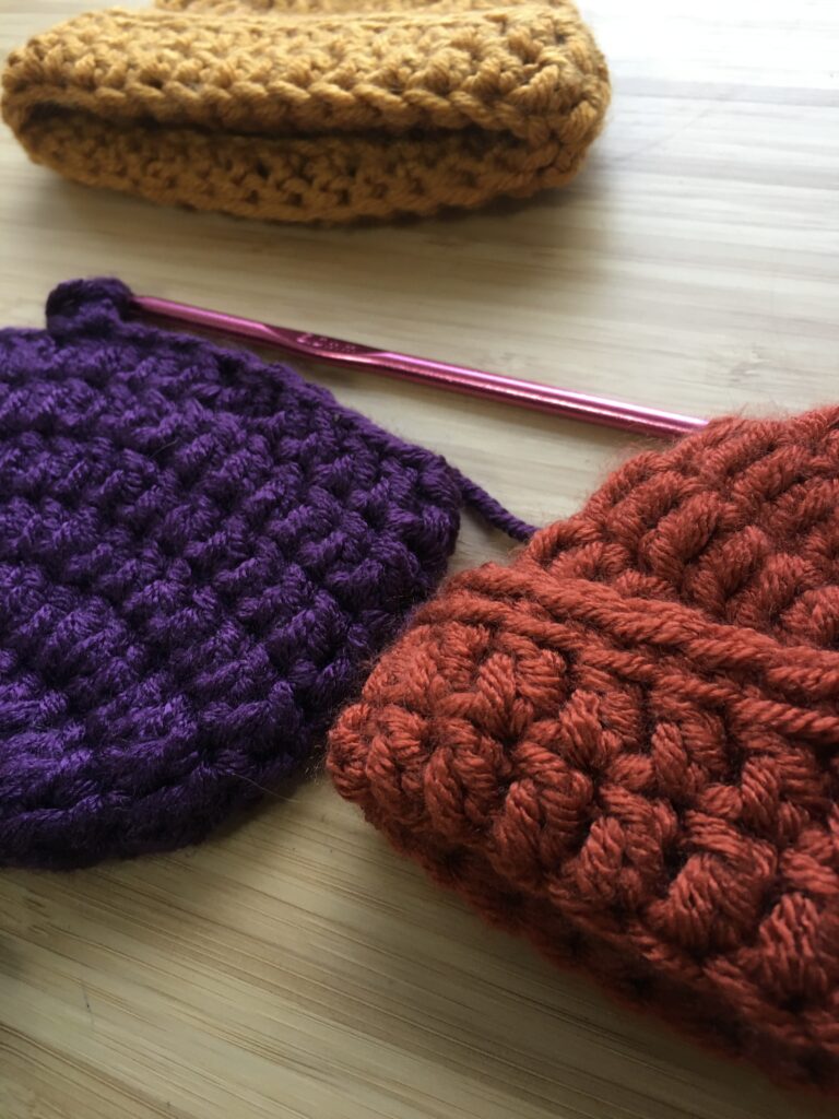 completed crochet newborn hat, a cap with a small brim in gold and orange resting on a light wooden background with an purple newborn hat resting in the middle on a pink crochet hook still being completed.