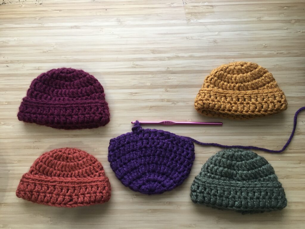 Four completed crochet hats for newborns rest on a wooden background, the hats are maroon, gold, green and burnt orange in colour, all four hats have their brim towards the bottom of the photo, an unfinished purple crochet hat rests in the opposite direction in the middle of the completed four hats on a pink aluminum crochet hook