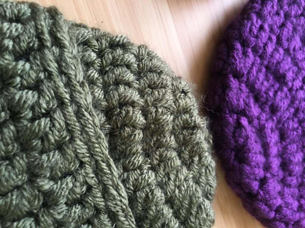 A completed green crochet hat is with a cap and brim is seen up close do that the double crochet stitches are recognizable, there is an incomplete purple hat resting in the opposite direction of the green hat (so they are mirrored images) both rest on a light wooden backdrop. 