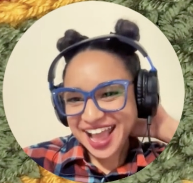 Lulu wears a big smile and big headphones in this frame taken from a crochet video tutorial. I am wearing an orange and blue plaid shirt and my classic blue glasses with symmetrical buns in my hair. I'm against beige backdrop in a circle. There is a background of green crochet around the circle acting as a frame.