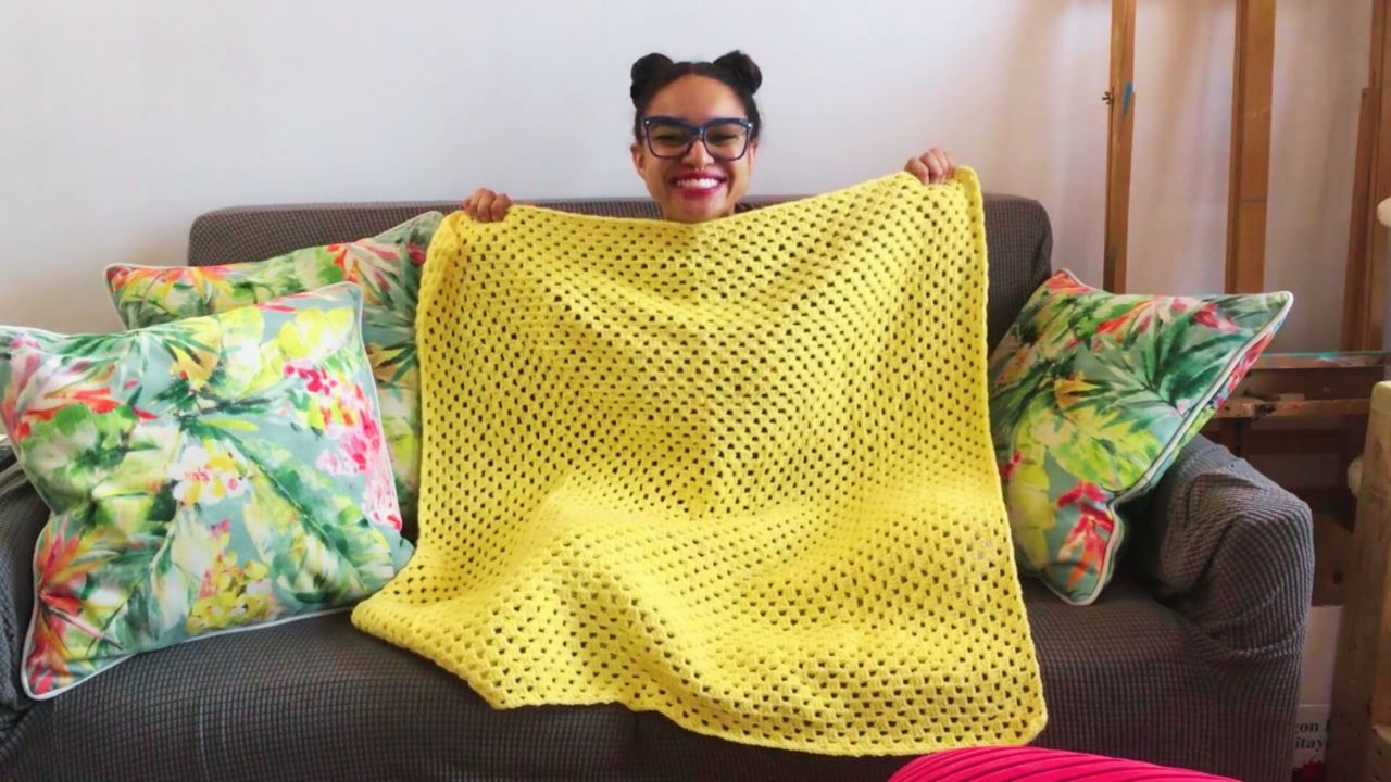 Lulu sits on a couch smiling with a large yellow granny square in front of her, green flowery pillows rest on either side