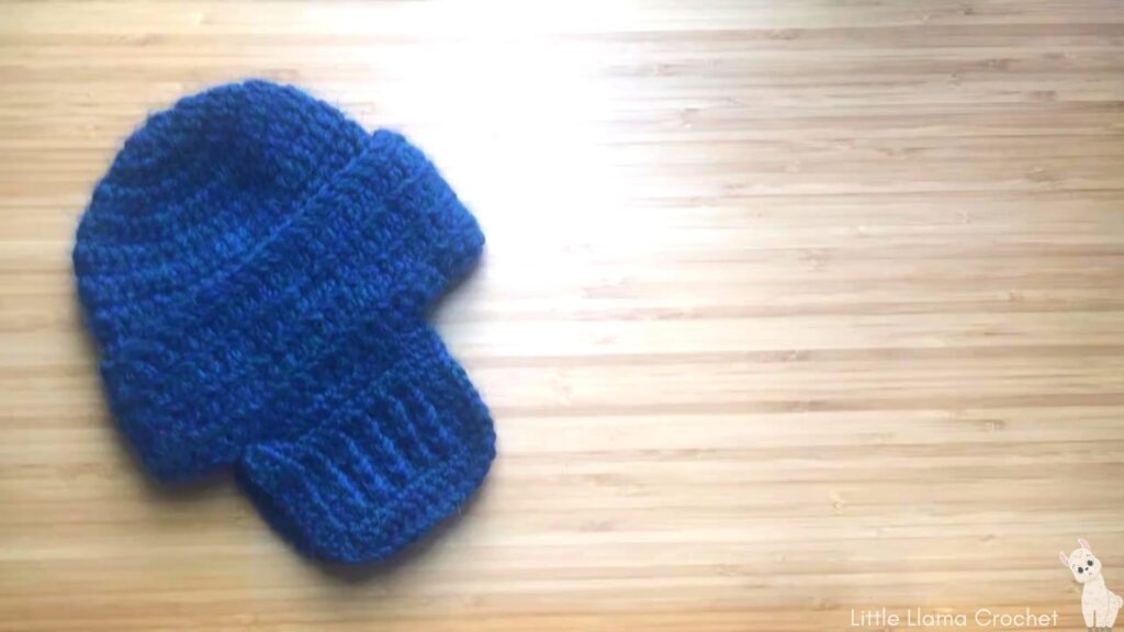 A flat overhead photo of a blue crochet baby hat with ear flaps lay on a wooden desk. The Blue hat has a cap, thick brim and ear flaps using a textured stitch. There is a cute little llama in the bottom right hand corner with the words. Little Llama Crochet written out.