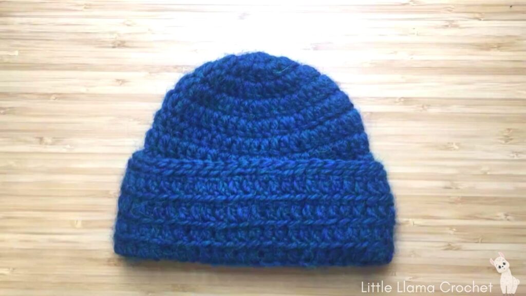 A flat overhead photo of a blue crochet baby cap with a thick brim lays on a wooden desk. There is a cute white little llama in the bottom right hand corner with the words. Little Llama Crochet written out.