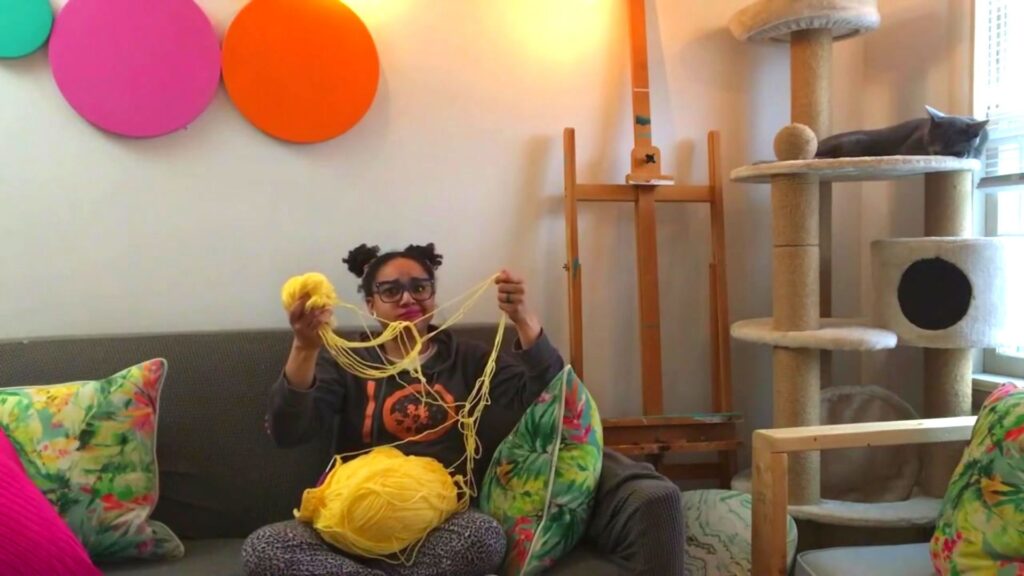 Lulu sits on a couch with a yellow one pound ball of yarn in her lap and I am holding up the largest yarn knot I have ever seen in my life. I do not look impressed. I am wearing a grey sweatshirt and pj pants. There are pink and orange circles on the wall behind me and a cat tree the right of the photo with a grey sleeping cat on the upper level.